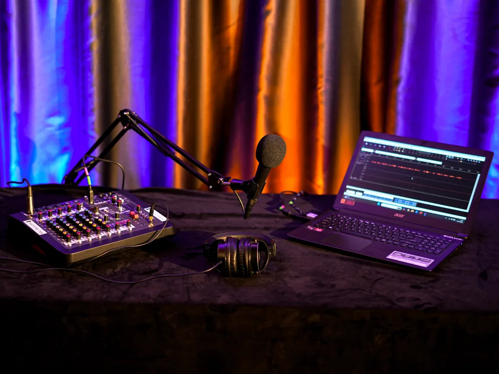 Audio podkāstu komplekts vienai personai ar mikrofonu, mikšerpulti, austiņām un datoru uz krāsaini izgaismota violeti oranža fona.