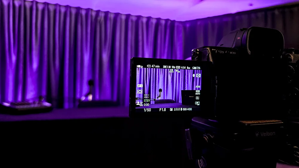 Video podkāstu studija vienai personai ar kameru priekšplānā un violetu fona apgaismojumu.