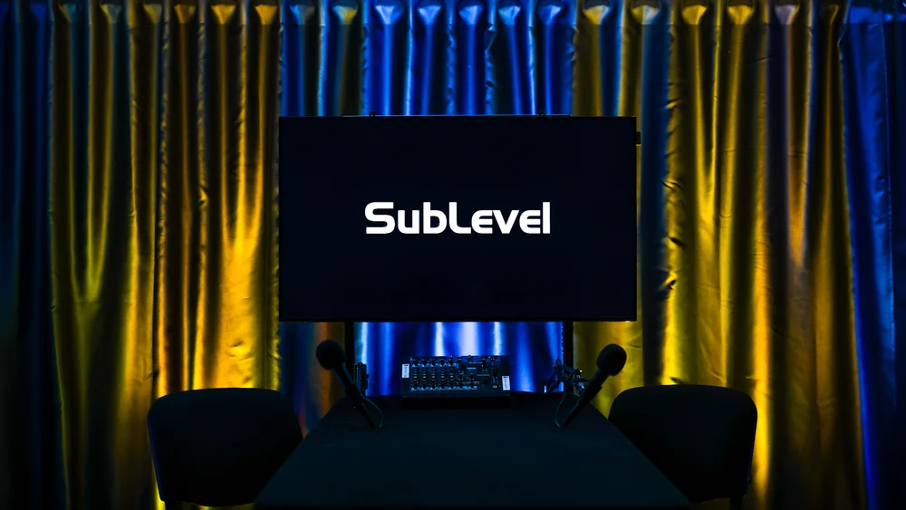 Pro studijas iekārtojums divām personām ar TV ekrānu, kas rāda 'SubLevel' logo, diviem mikrofoniem, mikšerpulti un kameru priekšplānā uz zili sarkana fona.
