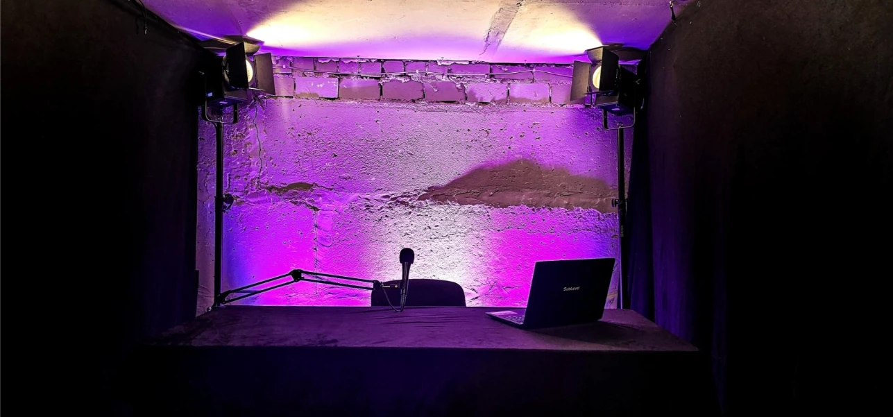 Video podkāsta studija vienai personai ar industriālu fonu, violetām un baltām LED gaismām, prožektoru un portatīvo datoru Rīgā